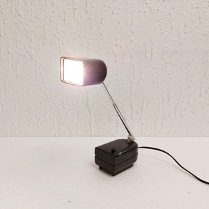 Puede incluir: Una lámpara de escritorio marrón con una pantalla rectangular y un brazo flexible. La lámpara tiene una base negra y un cable plateado.