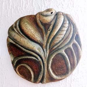 Puede incluir: Escultura mural redonda y texturizada con un diseño en forma de hoja. Presenta tonos marrones, beige y verdes, con una estructura central en forma de vena y formas redondeadas y abiertas. La obra de arte está montada en una pared blanca texturizada.