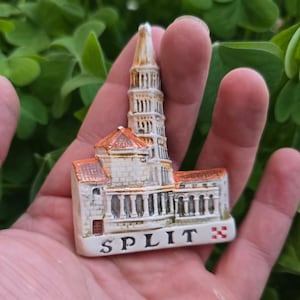 Könnte beinhalten: Ein Split-Souvenir, ein Miniaturmodell eines Gebäudes mit hohem Turm und rotem Dach. Das Wort "SPLIT" ist auf der Basis aufgedruckt. Das Souvenir wird in einer Hand vor einem Hintergrund aus grünem Laub gehalten.