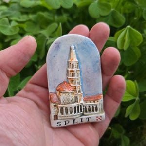 Könnte beinhalten: Ein Souvenir-Magnet aus Split, Kroatien, mit einer detaillierten Darstellung eines historischen Gebäudes mit hohem Glockenturm und rotem Ziegeldach. Der Magnet ist oben gewölbt und trägt unten das Wort "SPLIT".