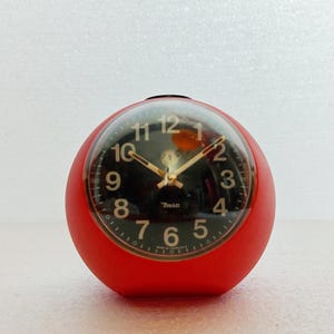 May include: A red, spherical alarm clock with a black face and gold hands. The clock face displays white numerals and the text "7min". The clock has a clear, rounded cover.