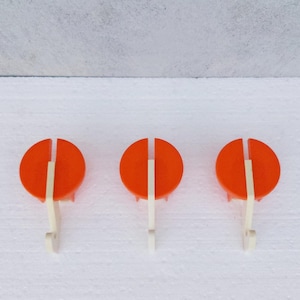 May include: Three orange and white wall hooks. Each hook features a circular orange top with a vertical white stripe and a white hook. The hooks are arranged in a row against a white background.