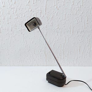 Puede incluir: Una lámpara de escritorio negra con una cabeza rectangular y un brazo largo y delgado de color plateado. La base de la lámpara también es negra y rectangular. La lámpara está enchufada a una toma de corriente con un cable negro. La lámpara está sobre una superficie blanca.