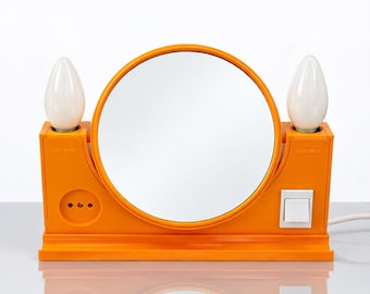 May include: A bright orange vanity mirror featuring a circular glass center flanked by two white light bulbs. The base includes a white power switch on the right and a circular outlet on the left, with the text MAX 25 W printed near each bulb socket.