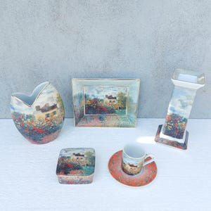 Op de afbeelding: Een verzameling keramische decoratieve items met een landschapsschilderij van een huis en kleurrijke bloemen. Inclusief een vaas, fotolijst, doos, kandelaar en kop en schotel. De items hebben een gouden rand.