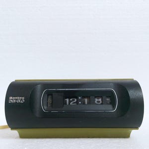 Puede incluir: Un reloj de solapas Sankyo DIGI-GLO negro y verde oliva con una pantalla rectangular horizontal que muestra la hora 12:18. El dispositivo presenta una carcasa central oscura con una base clara y un cable de alimentación en el lado izquierdo.