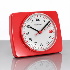 Op de afbeelding: Een rechthoekige rode Iskra Quarz bureauklok met een witte ronde wijzerplaat. De klok heeft zwarte Arabische cijfers, zwarte uren- en minutenwijzers, een rode secondewijzer en een klein datumvenster met het rode getal 15 op een witte achtergrond.