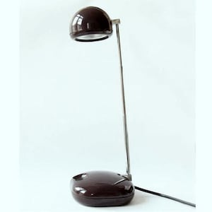 Könnte beinhalten: Eine dunkelbraune Schreibtischlampe mit rundem Schirm und Sockel, verbunden durch einen schlanken, silberfarbenen Arm. Die Lampe ist mit einem schwarzen Kabel an eine Steckdose angeschlossen. Das Design ist minimalistisch und modern.