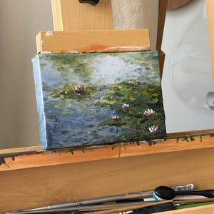 May include: A small rectangular canvas painting resting on a wooden easel. The artwork depicts a pond scene with green lily pads and small pink water lilies floating on blue and green water, rendered with visible, textured brushstrokes.