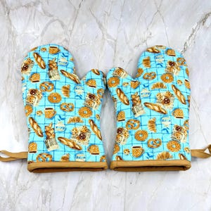 May include: A pair of quilted oven mitts featuring a light blue grid pattern with a repeating print of various baked goods including croissants, donuts, and coffee mugs. Each mitt has a solid tan fabric trim at the opening and a small loop.