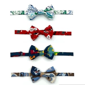 May include: Four colorful bow ties with various patterns are displayed against a white background. The bow ties feature designs including airplanes, dogs, and dinosaurs. Each bow tie has a matching adjustable neckband.