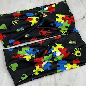 May include: Two black headbands with a colorful pattern of puzzle pieces, hearts, and handprints. The puzzle pieces are red, yellow, green, and blue. The headbands are made of a soft, stretchy fabric.