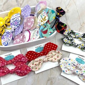 May include: A collection of handmade fabric hair bows in various patterns and colors. The bows include floral, polka dot, and sunflower designs. Some bows are displayed in a white oval dish, while others are on cards with the text "Home Stitch Design".