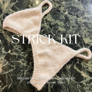Puede incluir: Un top de sujetador de punto beige con tirantes ajustables. El texto "STRICK-KIT" está impreso en blanco en la parte superior. El texto "Strickset fur deinen neuen Lieblings-BH in der Farbe deiner Wahl" está impreso debajo de la parte superior.