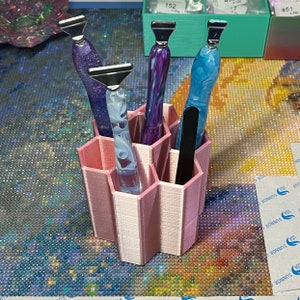 Pode incluir: Um organizador hexagonal rosa com seis compartimentos que cont&eacute;m quatro ferramentas com padr&otilde;es coloridos e em espiral. As ferramentas t&ecirc;m pontas de metal prateadas.