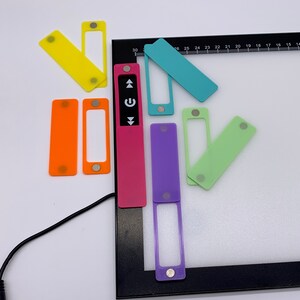 Op de afbeelding: Een set kleurrijke magnetische labels met een aan/uit-knop en pijltoetsen. De labels zijn bevestigd aan een zwart frame met een wit oppervlak. De labels zijn gemaakt van doorschijnend plastic en hebben een magnetische achterkant.