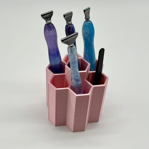 Pode incluir: Um organizador hexagonal rosa com seis compartimentos cont&eacute;m quatro ferramentas com al&ccedil;as azuis e roxas. O organizador &eacute; feito de pl&aacute;stico impresso em 3D.