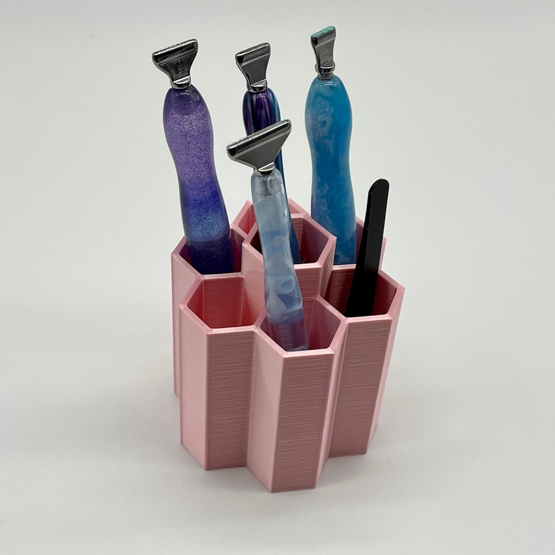 Pode incluir: Um organizador hexagonal rosa com seis compartimentos cont&eacute;m quatro ferramentas com al&ccedil;as azuis e roxas. O organizador &eacute; feito de pl&aacute;stico impresso em 3D.