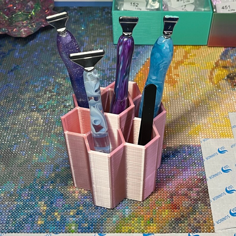 Pode incluir: Um organizador hexagonal rosa com seis compartimentos que cont&eacute;m quatro ferramentas com padr&otilde;es coloridos e em espiral. As ferramentas t&ecirc;m pontas de metal prateadas.