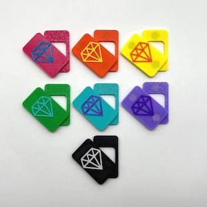 Op de afbeelding: Een verzameling kleurrijke, ruitvormige accessoires in verschillende kleuren, waaronder roze, oranje, geel, groen, turquoise, paars en zwart. Elk stuk heeft een ruitvormige afbeelding in een contrasterende kleur. De accessoires hebben een rechthoekige vorm met een uitsparing.