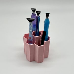 Pode incluir: Um organizador hexagonal rosa com quatro ranhuras para guardar ferramentas. O organizador tem uma ferramenta preta na ranhura central e tr&ecirc;s ferramentas coloridas nas outras ranhuras.