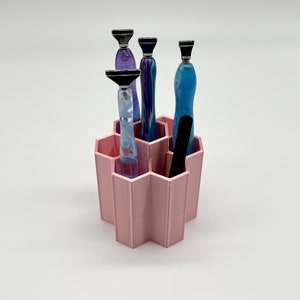 Pode incluir: Organizador hexagonal rosa com seis compartimentos, contendo quatro ferramentas de vidro com pontas prateadas e uma ferramenta preta. As ferramentas est&atilde;o dispostas verticalmente nos compartimentos.