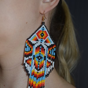 May include: A pair of long, beaded earrings with a geometric design in shades of red, orange, yellow, blue, black, and white. The earrings are made of small, colorful beads and have a dangling, fringe-like design.