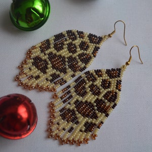 May include: A pair of long, beaded earrings with a brown and cream leopard print pattern. The earrings are made with small, round beads and have gold-coloured hooks.