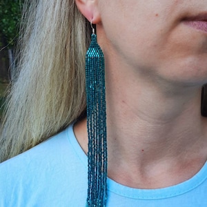 Puede incluir: Pendientes de borla de cuentas azul turquesa que cuelgan de la oreja de una persona. Los pendientes son largos y cuelgan hasta el cuello de la persona.