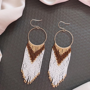 May include: A pair of long, beaded dangle earrings with gold hoops. The earrings feature white, brown, and gold beads in a cascading pattern.