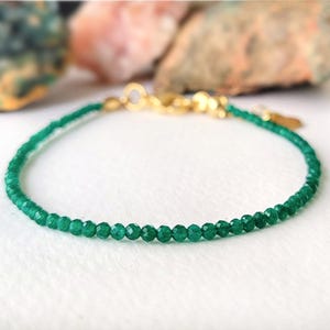Puede incluir: Una delicada pulsera de cuentas verdes con un cierre dorado. La pulsera presenta pequeñas cuentas verdes facetadas ensartadas. El cierre es de color dorado brillante. El fondo está borroso, con toques de verde y rosa.