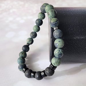 May include: A beaded bracelet featuring a mix of green, black, and gray stones. The bracelet includes round, patterned green beads, dark gray beads, and black lava rock beads, connected by small, silver-colored spacers. The bracelet is displayed on a white surface.