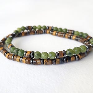 May include: Two beaded bracelets. One bracelet features olive green round beads and dark gray cylindrical beads. The other bracelet has brown rectangular beads and dark gray cylindrical beads. The bracelets are layered.