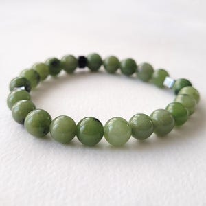 May include: A beaded bracelet featuring round, polished green jade stones. The bracelet is strung on an elastic band and includes a few small, silver-colored accent beads. The jade beads vary slightly in shade, creating a natural, organic look.