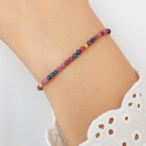 May include: Delicate beaded bracelet featuring small, round beads in shades of pink, blue, and gold. The bracelet is strung on a thin, flexible cord and is designed to be worn on the wrist. The beads are evenly spaced, creating a simple yet elegant design.