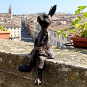 May include: A dark brown cast iron rabbit sculpture, perched on a stone ledge. The rabbit is depicted in a seated position, with its legs dangling over the edge. The sculpture has a textured surface and is set against a blurred cityscape background.