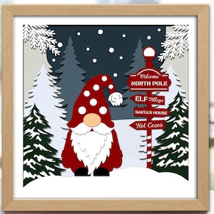 May include: A framed 3D layered paper cut shadow box featuring a Christmas gnome with a red hat and white beard. The scene includes a signpost pointing to the North Pole, Elf Village, Santa's House, and Hot Cocoa. The artwork is 20x20 cm.