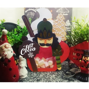 Puede incluir: Una taza de cartón roja y blanca para chocolate caliente con un diseño de cascanueces. La taza está llena de malvaviscos y tiene un letrero marrón que dice "Chocolate caliente" con un copo de nieve.