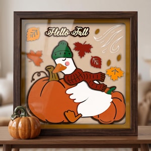 Può includere: Una cornice quadrata a cassetta contenente una scena artistica in carta a strati con un'oca bianca che indossa un berretto di lana verde e una sciarpa rossa, appoggiata su zucche arancioni. Il testo "Hello Fall" è in alto tra foglie autunnali.