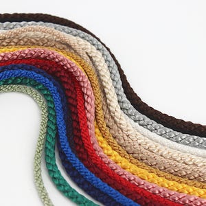 May include: A collection of braided cords arranged in a curved, fan-like pattern. The cords feature a smooth, satin-like finish and come in a variety of colors including dark brown, silver, beige, gold, pink, red, dark blue, and light green.