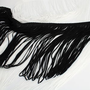 May include: A decorative trim featuring long, flowing fringe strands in contrasting black and white colors. The fringe is attached to a woven header band, creating a layered effect with the black section positioned over the white section.