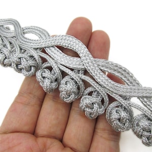 May include: Close-up of silver decorative trim with knotted accents. The trim features a braided design with small, round knots evenly spaced along the length. The metallic silver colour gives it a luxurious appearance, suitable for various craft projects.
