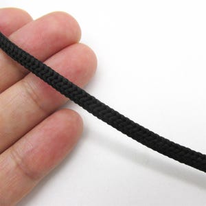May include: Close-up of a length of black braided cord. The cord is tightly woven, with a slightly textured surface. The background is plain white, highlighting the cord's dark color and detailed texture.