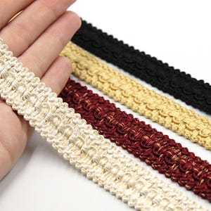 May include: Close-up of decorative trim in various colours. The trim is made of woven material with a scalloped edge. Colours include cream, burgundy, gold, and black. The trim is likely used for sewing, crafting, or embellishing clothing and accessories.