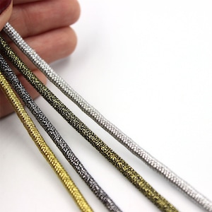 May include: Four strands of metallic cord in silver, gold, bronze, and black. The cords are lying on a white surface.