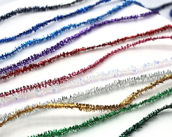May include: Assortment of colorful metallic tinsel trim, featuring shades of blue, purple, red, silver, gold, and green. The tinsel is arranged in rows on a white surface.