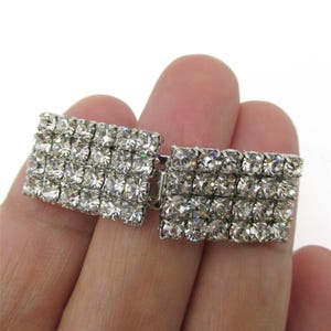 May include: A rectangular silver-tone metal clasp featuring three horizontal rows of clear, faceted rhinestones. The piece consists of two connected sections that form a continuous grid of sparkling stones, designed to function as a decorative fastener.