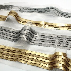 May include: Three strips of decorative trim featuring rows of metallic gold and silver rectangular sequins attached to a sheer white mesh fabric. The sequins are arranged in horizontal layers, creating a textured, shimmering appearance.