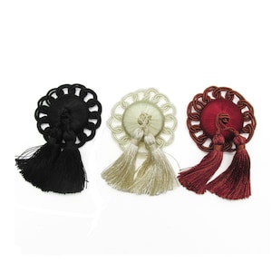 Op de afbeelding: Drie decoratieve ornamenten met een centrale ronde knop omwikkeld met satijndraad, omringd door een gevlochten koordrand en afgewerkt met twee hangende kwastjes. De set bevat een zwart, een crème en een donkerrood ornament.