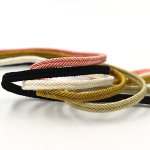 May include: A close-up of five different colours of braided cord. The cords are pink, black, white, gold, and beige. The cords are arranged in a pile, with the pink cord on top.